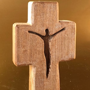 Op de afbeelding: Een houten kruis met een gegraveerd silhouet van een persoon aan de voorkant. Het kruis is lichtbruin van kleur en heeft een ruw, getextureerd oppervlak.