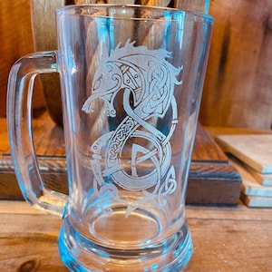May include: A clear glass beer stein with an etched design of a wolf's head with intricate knotwork and a runic symbol.