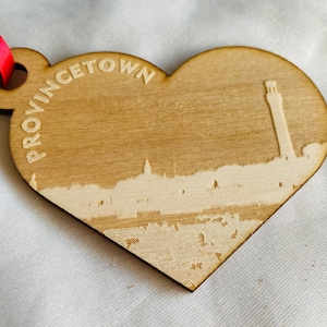 May include: A wooden heart-shaped ornament with the words "Provincetown" engraved on the top. The ornament features a white silhouette of the Provincetown skyline, including a lighthouse.