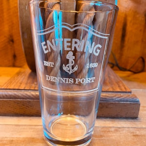 May include: A clear glass pint glass with the text "Entering Dennis Port Est 1689" etched on the side. The text is surrounded by a decorative border and an anchor symbol.