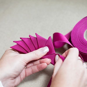 May include: A person's hands are shown folding a length of magenta ribbon into a fan shape. A spool of magenta ribbon is in the background.