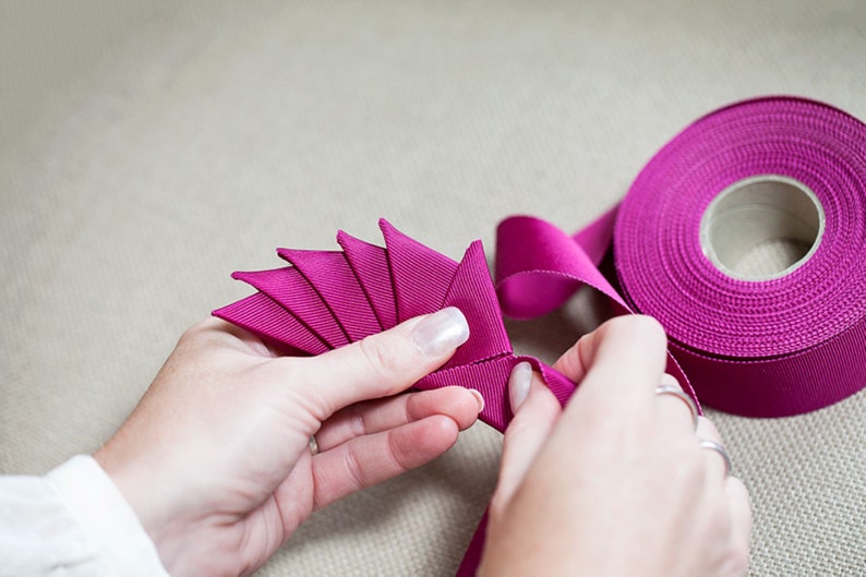 May include: A person's hands are shown folding a length of magenta ribbon into a fan shape. A spool of magenta ribbon is in the background.