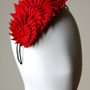 May include: A red fabric headband with a large, folded flower design. The headband is made of a soft, pliable material and is designed to be worn on the head.
