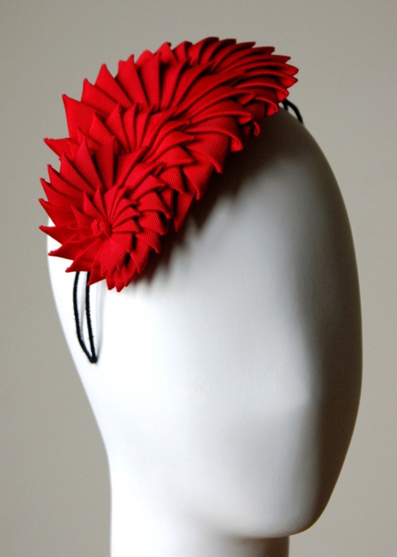 May include: A red fabric headband with a large, folded flower design. The headband is made of a soft, pliable material and is designed to be worn on the head.