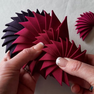 May include: A close-up of a hand holding a burgundy fabric flower made of multiple folded triangles. The flower is being made by hand and is in the process of being assembled. There is a smaller burgundy fabric flower in the background.