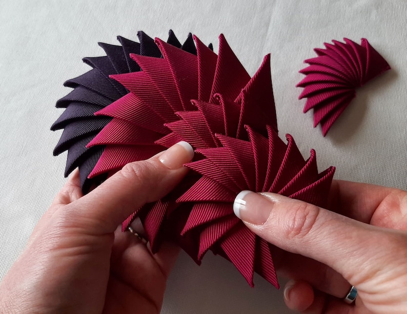 May include: A close-up of a hand holding a burgundy fabric flower made of multiple folded triangles. The flower is being made by hand and is in the process of being assembled. There is a smaller burgundy fabric flower in the background.