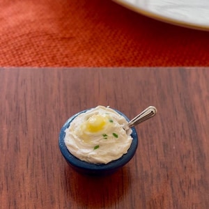 Puede incluir: Un pequeño tazón azul lleno de puré de patatas cubierto con una porción de mantequilla y cebollino. Una cuchara de plata descansa en el borde del tazón.