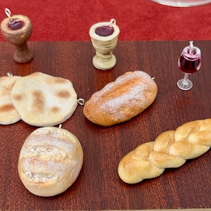 Może przedstawiać: Miniaturowe zawieszki z jedzeniem, w tym dwa podpłomyki, bochenek chleba, plecionka, okrągły bochenek i dwa kielichy z czerwoną cieczą. Obecna jest również mała szklanka czerwonego wina. Ozdoby znajdują się na ciemnobrązowej powierzchni.