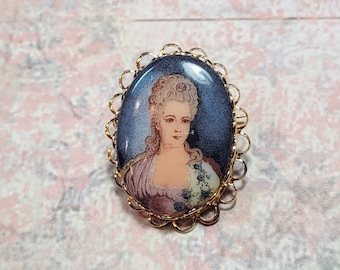 May include: A gold-toned brooch with a cameo featuring a woman in a white dress with a floral design. The cameo is set in a gold-toned oval frame with a decorative border.