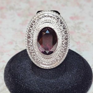 May include: An oval-shaped silver ring with an ornate filigree design. The ring features a large, deep purple gemstone at its center. The ring is displayed on a black velvet stand against a floral background.