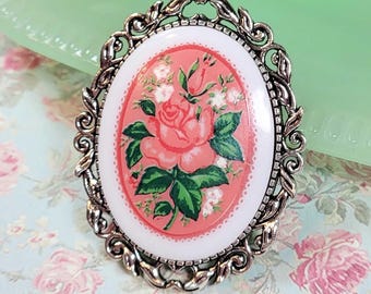 May include: A silver oval-shaped brooch with a pink and white floral design. The brooch features a large pink rose with green leaves and white flowers.