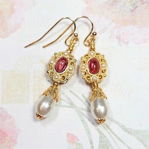 May include: A pair of gold-tone drop earrings featuring a central oval pink stone surrounded by small white pearls, with a larger white teardrop-shaped pearl dangling from the bottom of each piece, suspended from gold-tone ear wire hooks.