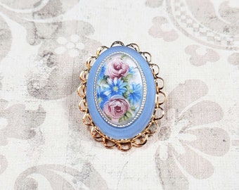 May include: Oval-shaped brooch with a light blue background and a floral design. The brooch features pink roses and blue flowers within a gold-colored filigree frame. A vintage-style accessory.