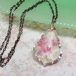 May include: A delicate necklace with a clear glass pendant featuring a floral pattern in shades of pink and green. The pendant is attached to a black chain.