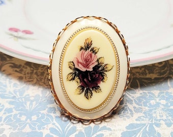 May include: Oval-shaped vintage brooch with a cream-colored background and a floral design featuring pink and burgundy roses. The brooch has a gold-tone filigree frame and a beaded border, perfect for adding a touch of vintage charm to any outfit.