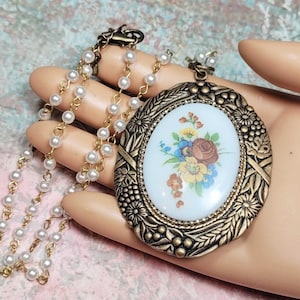 May include: Vintage-style necklace featuring a large oval pendant with a floral design in shades of red, blue, and yellow. The pendant is framed in a gold-toned metal with intricate detailing. The necklace has a pearl and gold-tone chain.