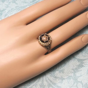 May include: A gold-toned ring with a black enamel face and a central pearl. The ring features intricate floral and geometric designs. The pearl is surrounded by a sunburst pattern. The ring is set on a light-colored background.
