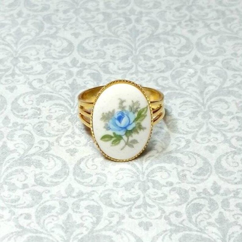 Pu&ograve; includere: Anello dorato con un elemento centrale ovale color crema con un disegno di rosa blu. L'anello ha un design a tripla fascia e un bordo dorato strutturato attorno all'immagine floreale. L'anello &egrave; posto su uno sfondo a motivi.