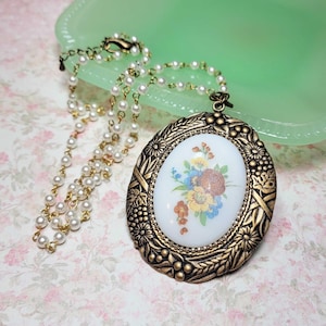 May include: A vintage-style necklace featuring a gold-toned oval pendant with a floral design in the center. The pendant is surrounded by an ornate frame and is suspended from a delicate pearl and gold chain. The necklace rests on a floral patterned surface.