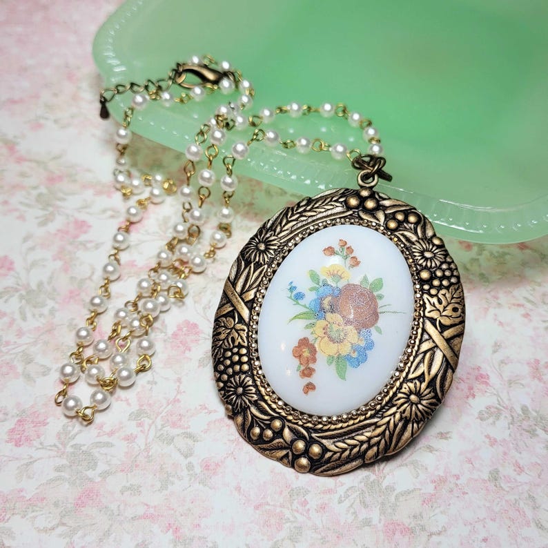 May include: A vintage-style necklace featuring a gold-toned oval pendant with a floral design in the center. The pendant is surrounded by an ornate frame and is suspended from a delicate pearl and gold chain. The necklace rests on a floral patterned surface.