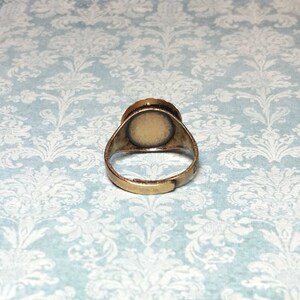 May include: A vintage-style adjustable ring with a brass-colored band. The ring features a circular setting with a light-colored surface, possibly for a cameo or other embellishment. The ring is set against a light blue background.