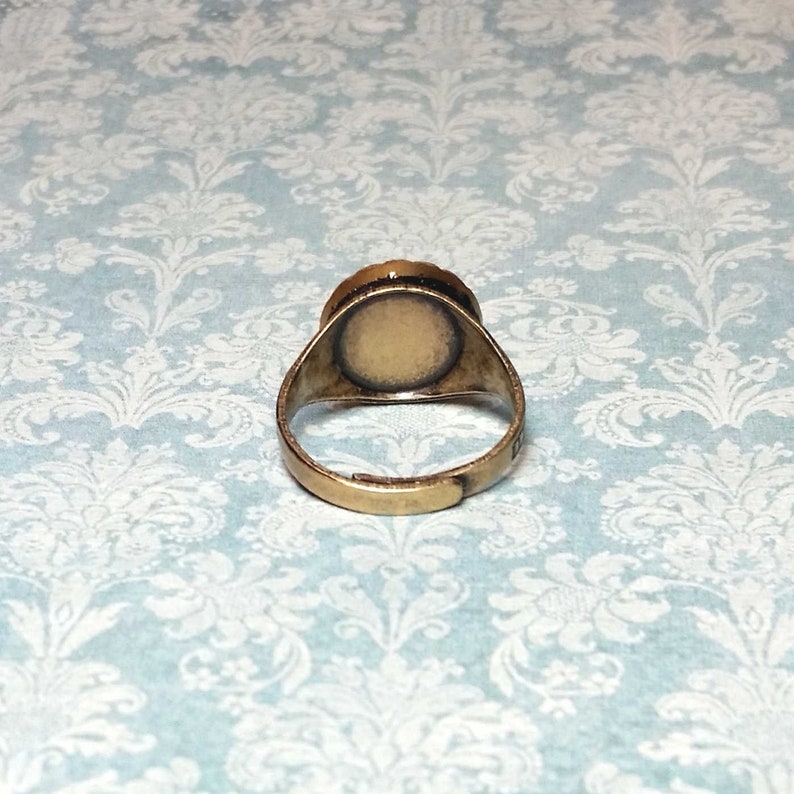 May include: A vintage-style adjustable ring with a brass-colored band. The ring features a circular setting with a light-colored surface, possibly for a cameo or other embellishment. The ring is set against a light blue background.