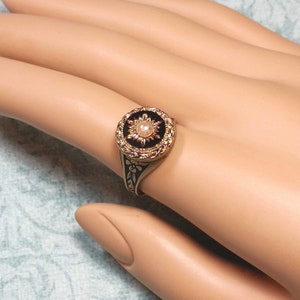 May include: A vintage-style ring with a gold-tone band and a black and gold circular face. The face features a central pearl-like stone surrounded by a sunburst design. The band has a floral pattern.