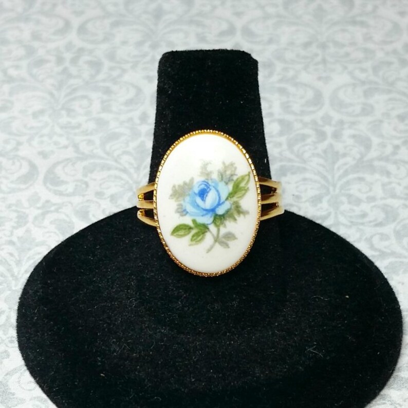 Pu&ograve; includere: Anello dorato con un elemento centrale ovale in porcellana con un motivo a rosa blu dipinto a mano. L'anello ha un design a tripla fascia e uno sfondo bianco. L'anello &egrave; esposto su un supporto nero.