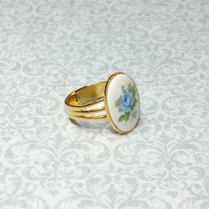 Pu&ograve; includere: Anello regolabile color oro con una faccia ovale in porcellana. La faccia mostra una rosa blu con foglie verdi su sfondo bianco, incorniciata da un bordo dorato perlato. L'anello ha un design a tripla fascia.