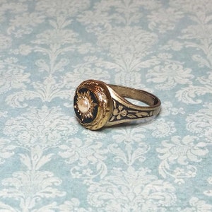 May include: A gold-toned ring with a black center featuring a pearl-like stone. The ring has intricate floral and sunburst designs. The band has floral engravings. The ring is set against a light blue background.
