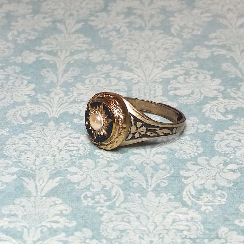 May include: A gold-toned ring with a black center featuring a pearl-like stone. The ring has intricate floral and sunburst designs. The band has floral engravings. The ring is set against a light blue background.