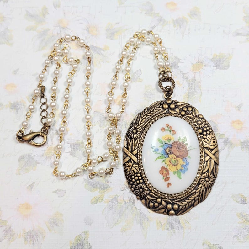 May include: A vintage-style necklace featuring a gold-toned pendant with an oval, floral-patterned center. The pendant is suspended from a delicate pearl and gold-tone chain. The necklace has a lobster clasp closure.