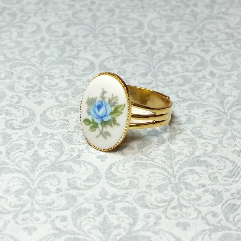 Pu&ograve; includere: Anello regolabile color oro con una faccia ovale in porcellana. La faccia mostra una rosa blu dipinta a mano con foglie verdi su sfondo bianco. L'anello ha un bordo dorato strutturato e un design a tre bande.