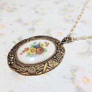 May include: Oval pendant necklace with a floral design. The pendant has a white background with a painted bouquet of flowers in shades of yellow, blue, and brown. The pendant is framed in a gold-colored metal with intricate detailing, and hangs from a delicate chain.
