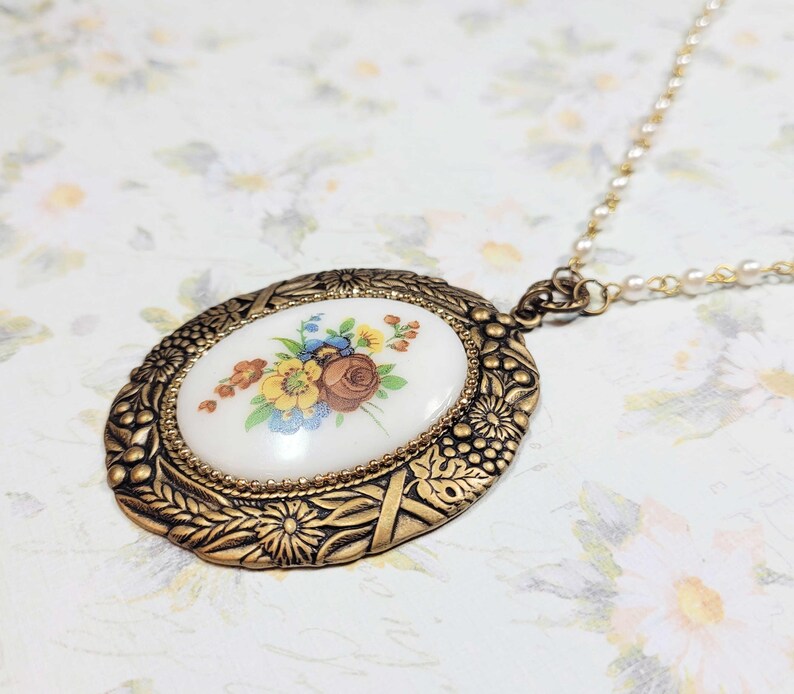 May include: Oval pendant necklace with a floral design. The pendant has a white background with a painted bouquet of flowers in shades of yellow, blue, and brown. The pendant is framed in a gold-colored metal with intricate detailing, and hangs from a delicate chain.