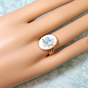 Pu&ograve; includere: Anello dorato con un viso ovale in ceramica bianca con un disegno di rosa blu. L'anello ha un'estetica floreale delicata, con una sottile fascia dorata e pi&ugrave; fasce di supporto. L'anello &egrave; mostrato su una mano di manichino.