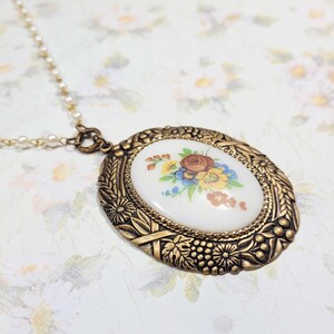 May include: A vintage gold-toned pendant necklace with an oval floral design. The pendant features a white center with a colorful flower bouquet. The ornate frame has a detailed, textured design. The necklace chain is a delicate strand of small pearls.