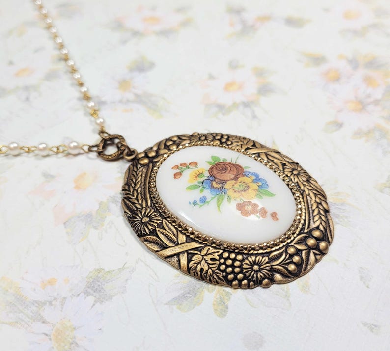 May include: A vintage gold-toned pendant necklace with an oval floral design. The pendant features a white center with a colorful flower bouquet. The ornate frame has a detailed, textured design. The necklace chain is a delicate strand of small pearls.