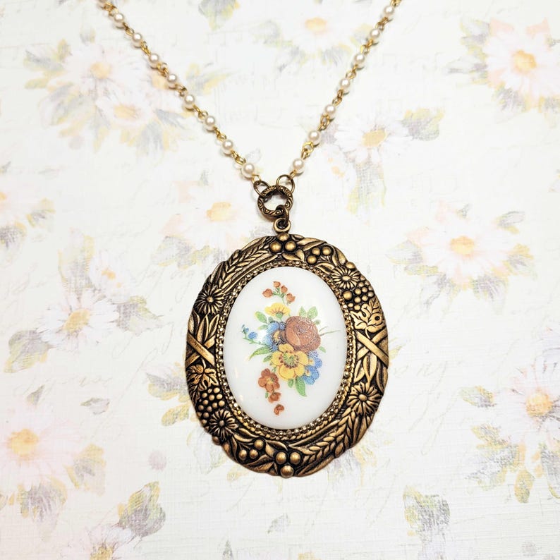 May include: A vintage-style necklace featuring an oval pendant with a floral design. The pendant has a gold-toned frame with intricate detailing. The chain is adorned with small, pearl-like beads, creating an elegant and classic look.