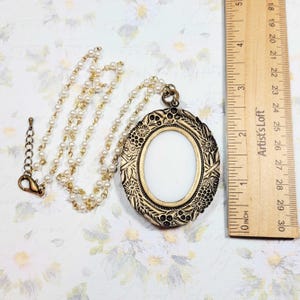 May include: An antique-style necklace with a gold-colored oval pendant and a pearl chain. The pendant has an ornate floral design around a white oval center. A wooden ruler is next to the necklace, measuring up to 12 inches.