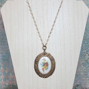May include: A vintage-style necklace featuring a large oval pendant with a floral design. The pendant is framed in a bronze-colored metal with intricate detailing. The necklace chain is composed of small, white beads.