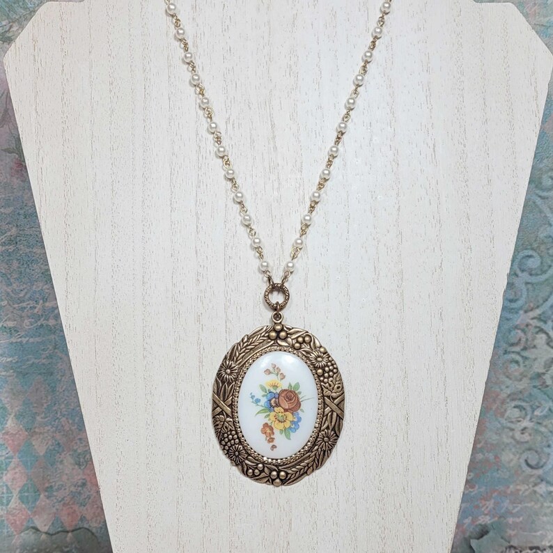 May include: A vintage-style necklace featuring a large oval pendant with a floral design. The pendant is framed in a bronze-colored metal with intricate detailing. The necklace chain is composed of small, white beads.