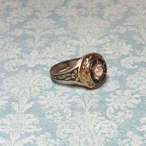 May include: A gold-toned ring with a black center and a pearl-like stone. The ring features intricate floral and sunburst designs. The band has a floral pattern. The ring is set against a light blue background.