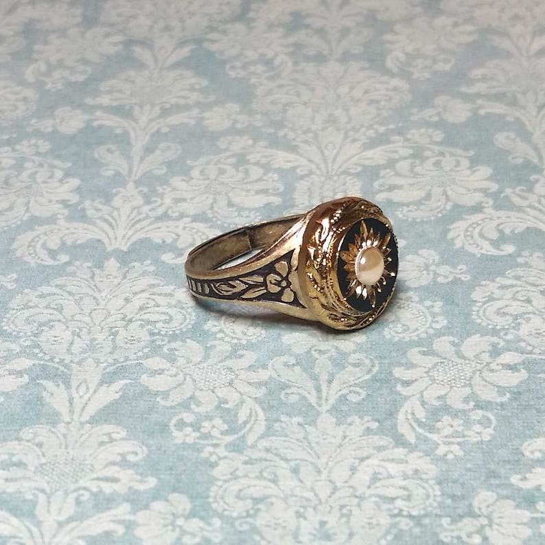 May include: A gold-toned ring with a black center and a pearl-like stone. The ring features intricate floral and sunburst designs. The band has a floral pattern. The ring is set against a light blue background.