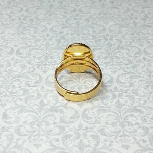 Pu&ograve; includere: Un anello regolabile color oro con un centro decorativo rotondo. L'anello presenta una fascia strutturata e un design circolare sulla parte superiore. L'anello &egrave; posto su uno sfondo a motivi.