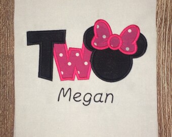 May include: A white fabric square featuring an embroidered applique design. The word Two is stitched in black and pink with white polka dots. A black mouse ear silhouette with a pink polka-dot bow is attached. The name Megan is stitched below.