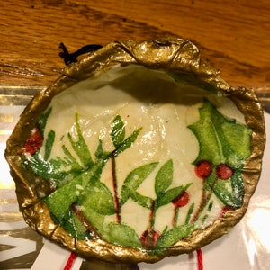 May include: A round, gold-rimmed ornament with a white background and a green and red holly pattern. The ornament has a red string attached to it.