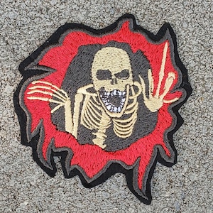 May include: Embroidered patch featuring a skeletal figure with a wide open mouth and outstretched hands. The skeleton is beige, set against a black background, framed by a red, flame-like border, and a black outline.