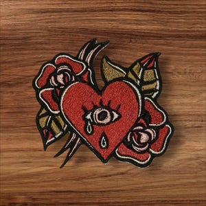 May include: Red embroidered heart with a single eye and a tear, surrounded by two pink and red roses with gold leaves.