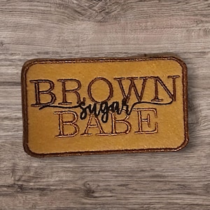 May include: A rectangular patch with the words "BROWN sugar BABE" embroidered in brown thread on a tan background. The patch has a brown border and is set against a wooden surface.